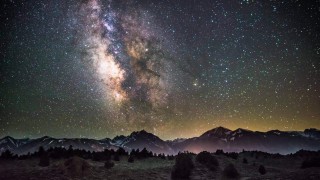 The milky way above a foggy landscape with mountains in the background and a forest in the foreground.